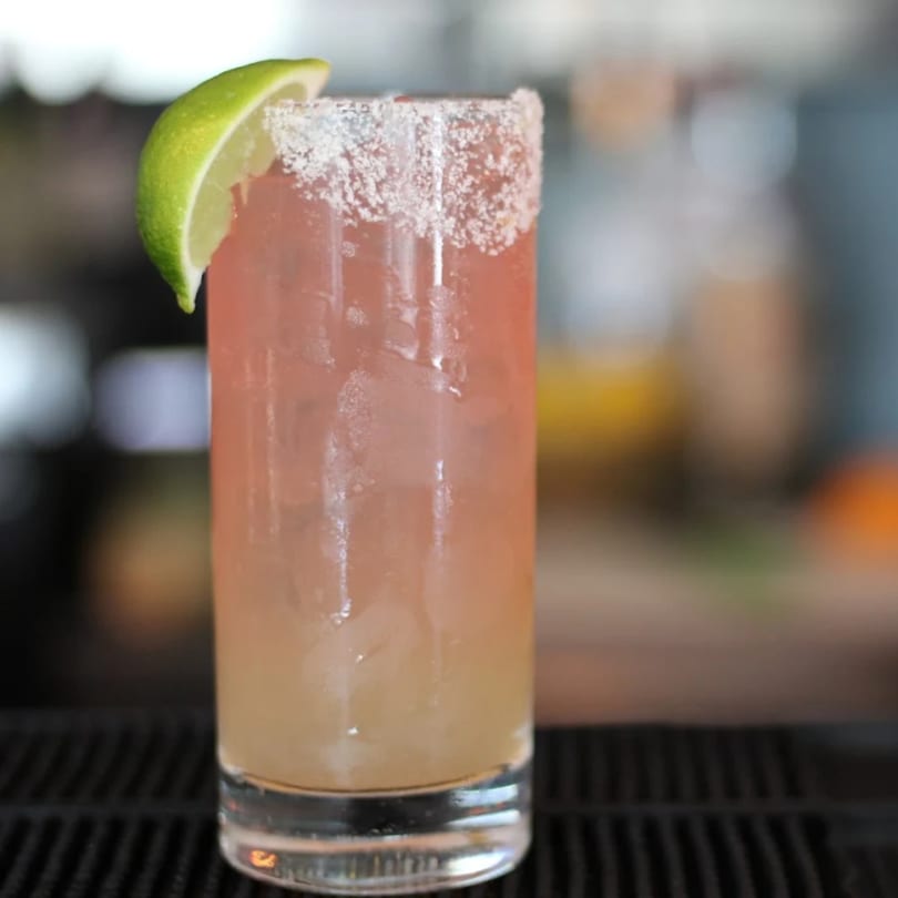 Pink cocktail with a salted rim and a lime wedge, on a dark bar surface.