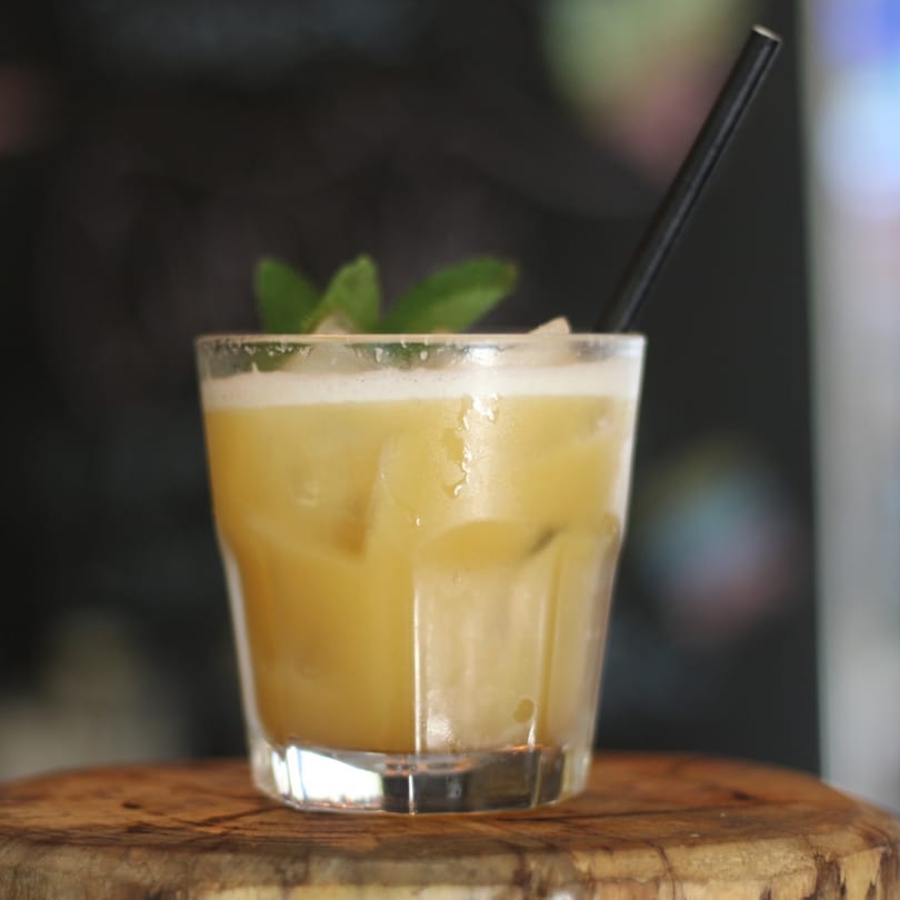 A Pinch Kitchen+Bar cocktail, yellow with foam, ice, mint, and a black straw on a wooden surface.