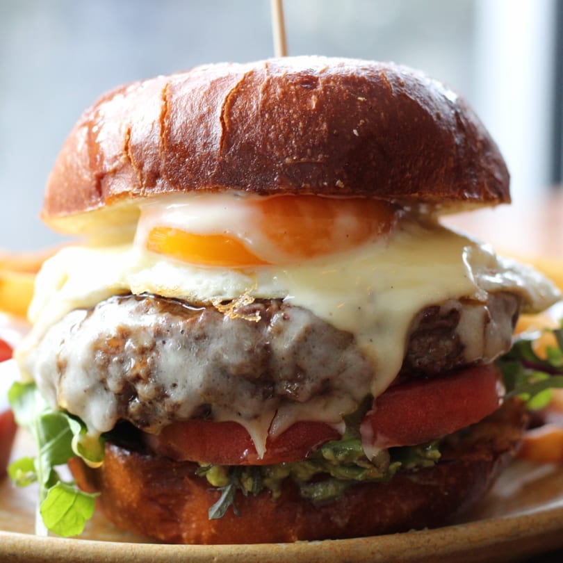 Close-up of a Pinch Kitchen+Bar burger with a fried egg, melted cheese, tomato, and greens.