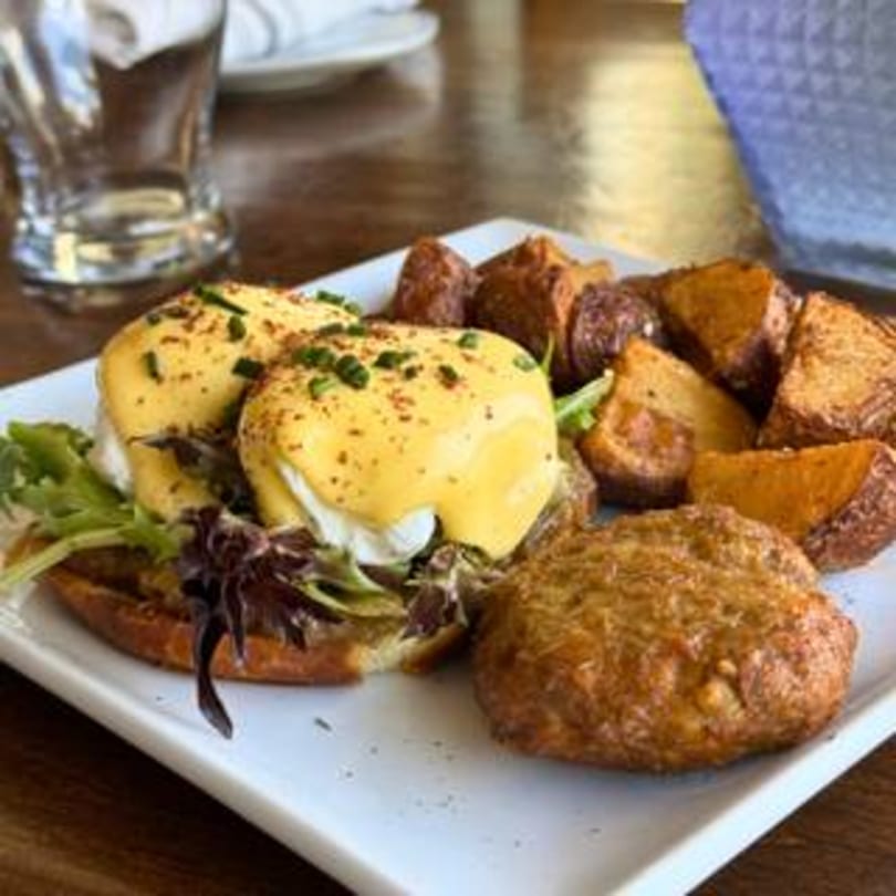 Eggs Benedict with hollandaise sauce, green onions, roasted potatoes, and a crab cake on a white plate.