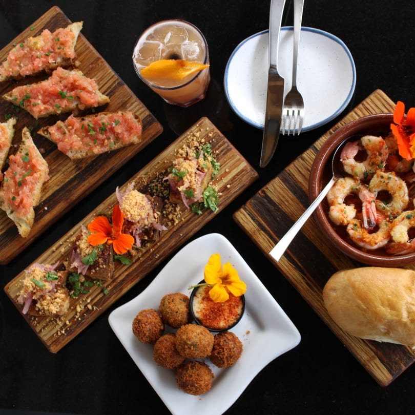 An overhead view of Pinch Kitchen+Bar's American tapas, including bruschetta, shrimp, croquettes, and a cocktail.