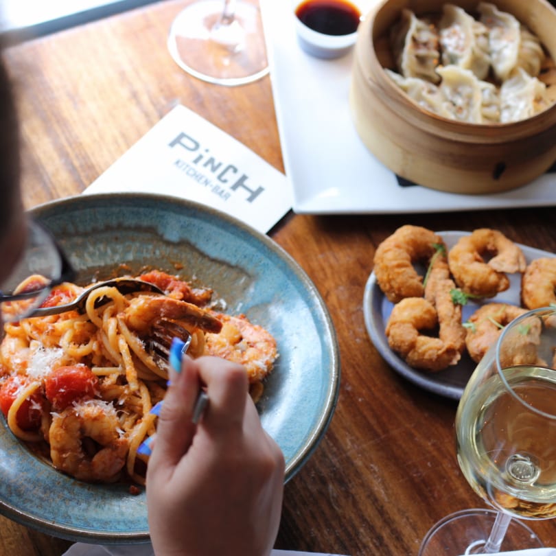 Diner eating shrimp pasta with fried shrimp, dumplings, and white wine on a wooden table at Pinch Kitchen+Bar.