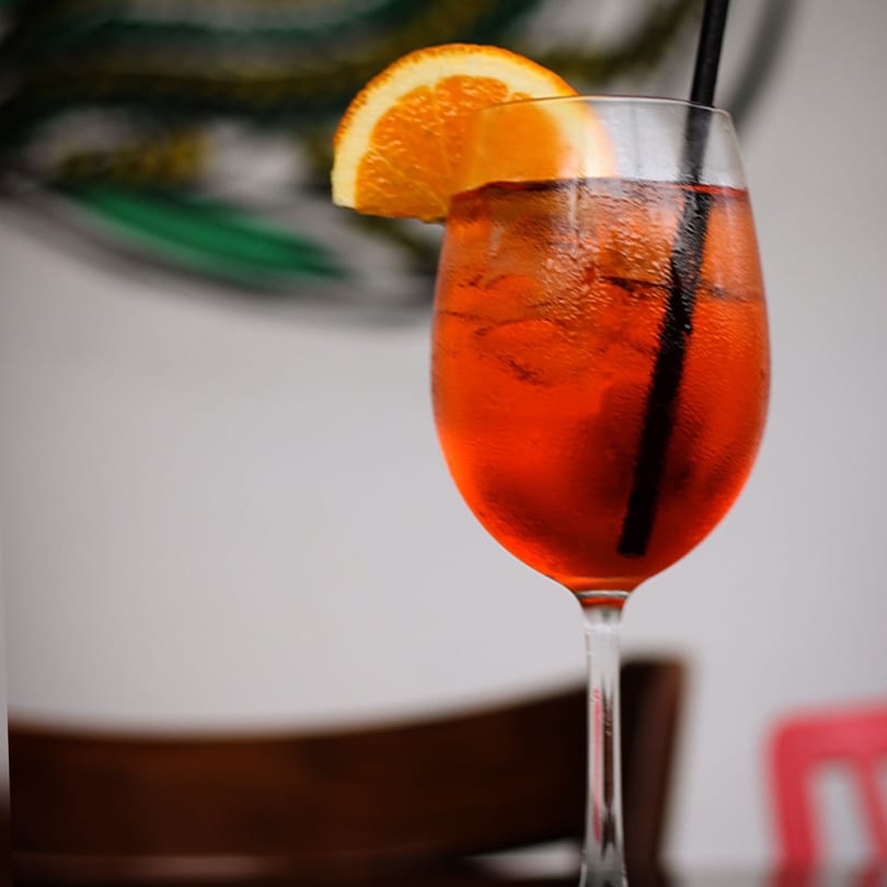 A vibrant orange cocktail with ice and an orange slice, served in a wine glass.