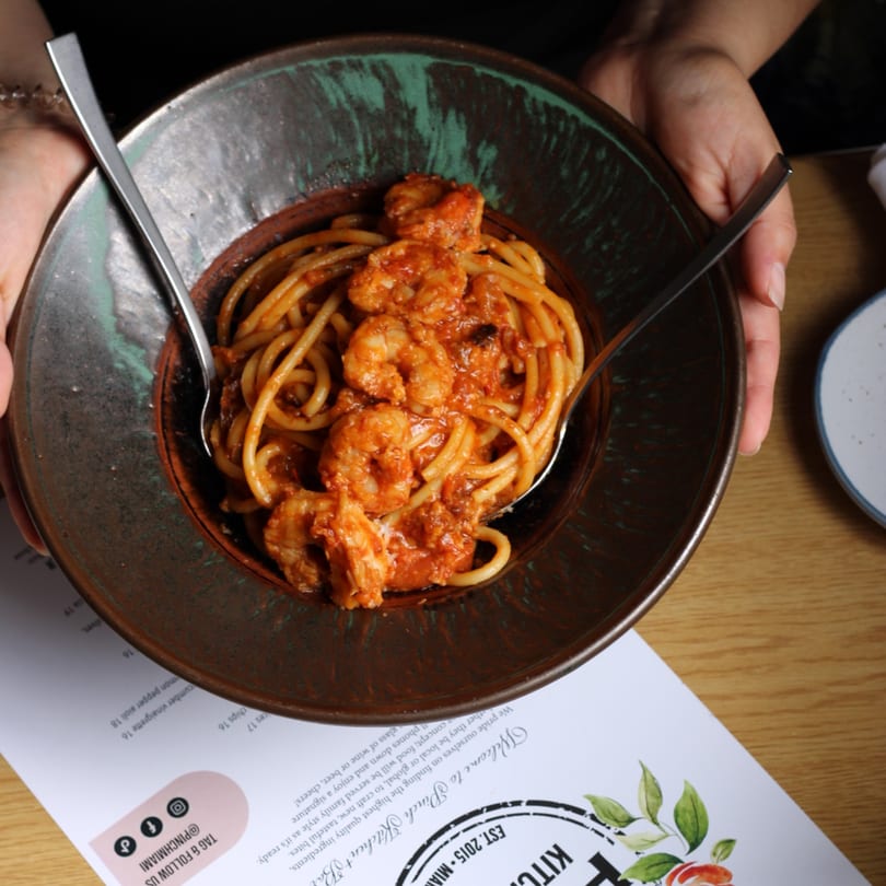 Hands holding a dark bowl of Pinch Kitchen+Bar's shrimp pasta with red sauce and two spoons.