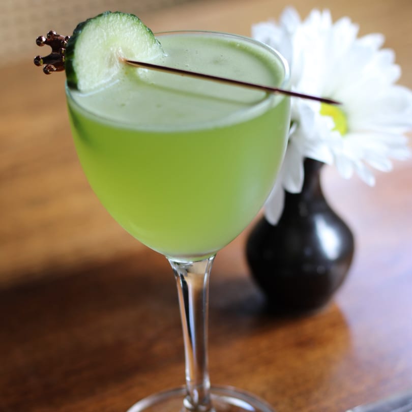 A vibrant green cocktail garnished with a cucumber slice and a decorative pick, next to a white flower.