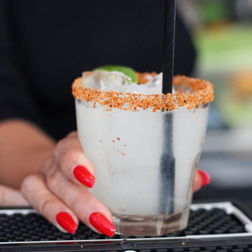 A hand with red nails holds a Pinch Kitchen+Bar margarita with a chili-salted rim and lime.