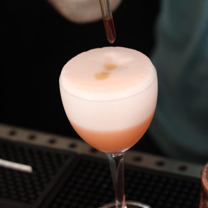 A bartender adds drops of bitters to a foamy pink cocktail in a coupe glass at Pinch Kitchen+Bar.